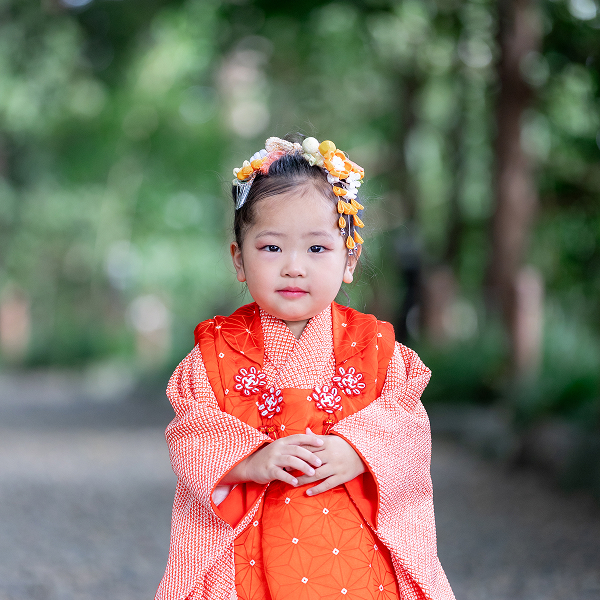神社の木々を背景に朱色の着物にオレンジの髪飾りをつけた幼い女の子