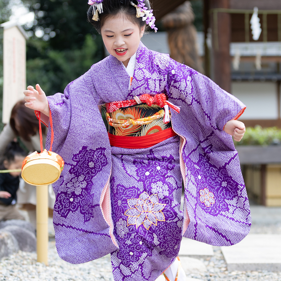神社にて紫の着物を着た女の子が七五三の小物を持ちながら駆け寄ってくる様子