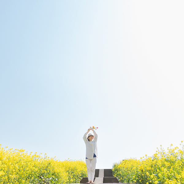 青空の下に菜の花が広がる中心に女性が手をあげて清々しく立っている