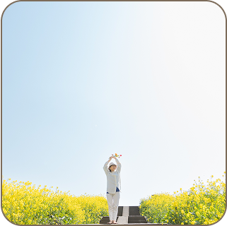 青空の下に菜の花が広がる中心に女性が手をあげて清々しく立っている
