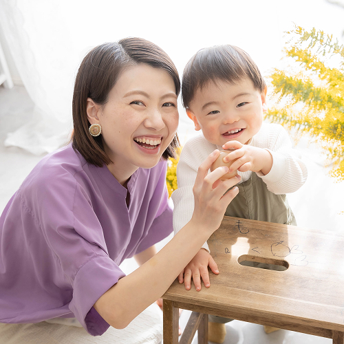 ミモザの花を背景にママと男の子が同じおもちゃを一緒に左右から持ちながら笑顔を向けている