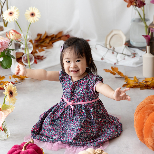 秋のお花やカボチャで装飾されたスタジオで、濃い色合いの細かい花柄のワンピースを着た女の子が笑って両手を広げている