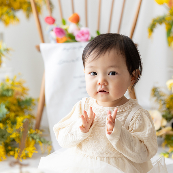 ミモザやチューリップ等の春のお花で彩られたスタジオにて、1歳のケーキに向かって座り手を叩く仕草をしている白いドレスを着た女の子の赤ちゃん