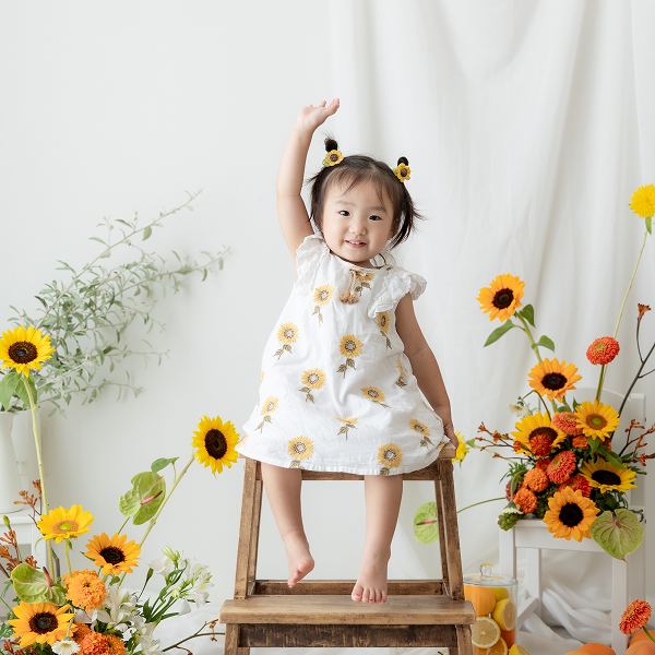 向日葵撮影会のスタジオの木の椅子に座り、元気よく右手をあげている幼い女の子
