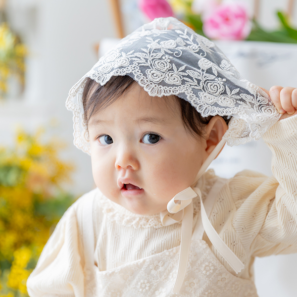 ミモザやチューリップなど春のお花を背景に女の子の赤ちゃんがお座りして帽子のツバをあげる仕草をしながらこちらを見ている