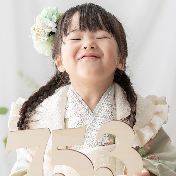 淡いグリーンの着物を着た女の子が七五三の木の装飾プレートを正面に持ち、少し甘えた満遍の笑みをこらえている瞬間