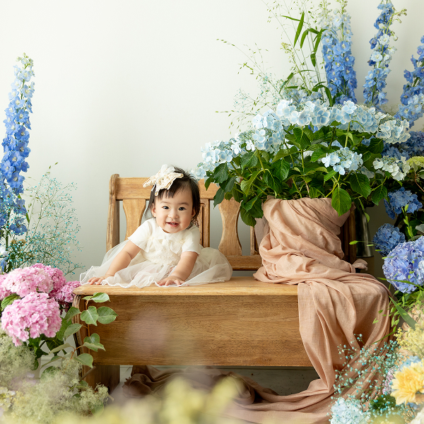 紫陽花に彩られたスタジオの木のベンチにお座りする、白いドレスと白い髪飾りを纏った笑顔の赤ちゃん
